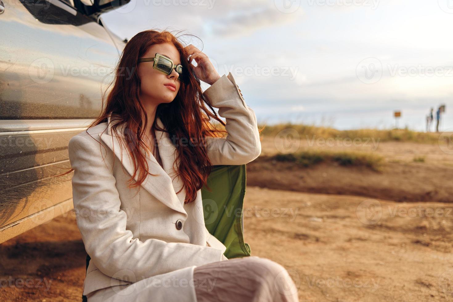 bonita mulher sentado em uma cadeira dentro uma campo vestindo Sombrio óculos perto a carro ensolarado dia foto