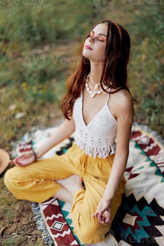 uma jovem hippie mulher medita dentro natureza dentro a parque, sentado dentro uma lótus posição em dela colorida xadrez e desfrutando harmonia com a mundo dentro roupas ecológicas foto