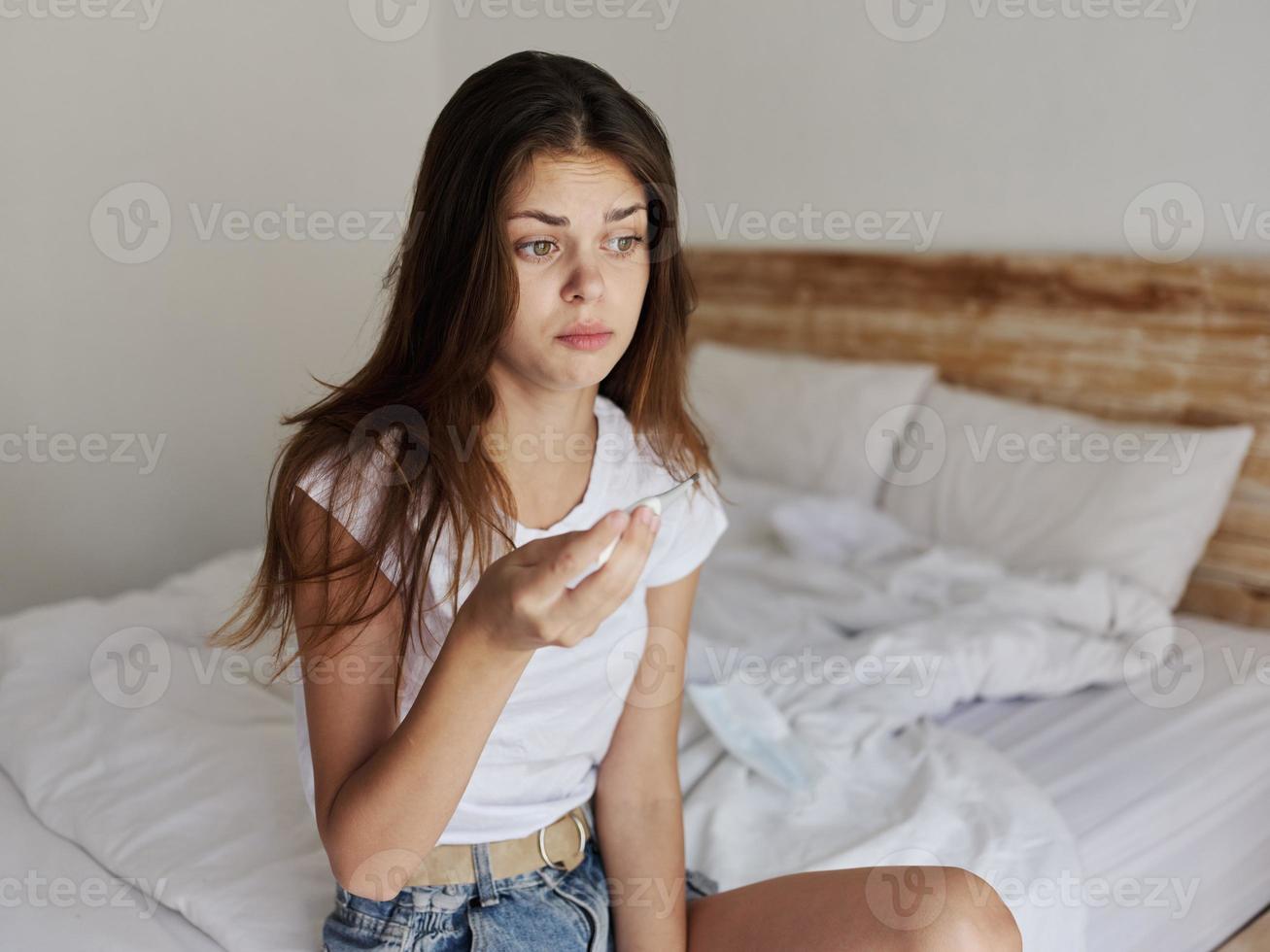 uma mulher senta em uma cama dentro uma hotel com uma termômetro dentro dela mãos foto