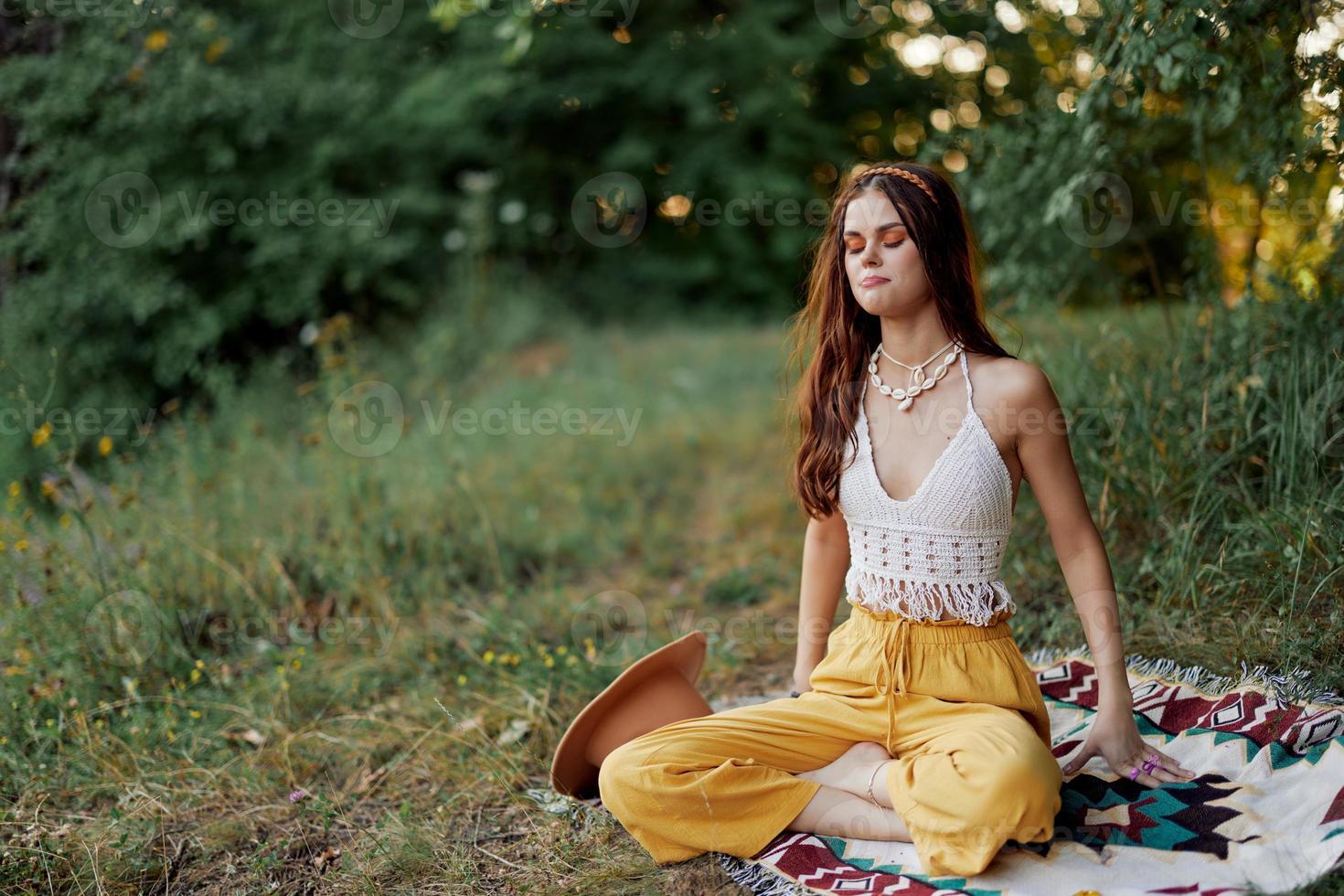 uma jovem hippie mulher medita dentro natureza dentro a parque, sentado dentro uma lótus posição em dela colorida xadrez e desfrutando harmonia com a mundo dentro roupas ecológicas foto