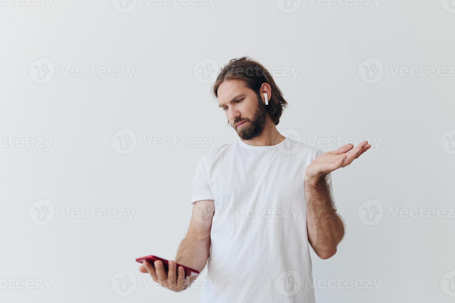 uma homem com uma barba blogueiro dentro uma branco camiseta com uma telefone e sem fio fones de ouvido parece para dentro a telefone e spreads dele mãos para a lado em uma branco fundo isolado cópia de espaço foto