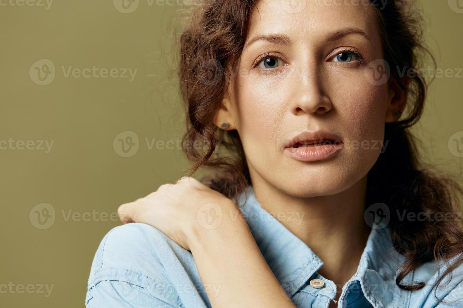 fechar-se retrato do bonita encaracolado lindo senhora dentro jeans casual camisa abraços ela mesma posando isolado em sobre Oliva verde pastel fundo. ser você mesmo. pessoas estilo de vida emoções conceito. cópia de espaço foto