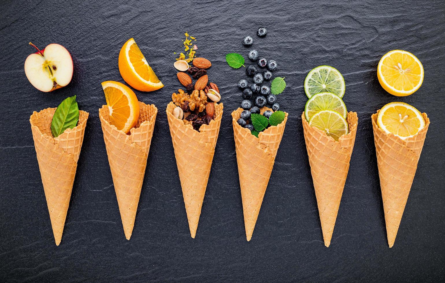 vários ingredientes para sabores de sorvete em cones mostrando mirtilos, limão, pistache, amêndoas, laranja, chocolate, baunilha e café em um fundo de pedra escura. verão e o conceito de menu doce. foto