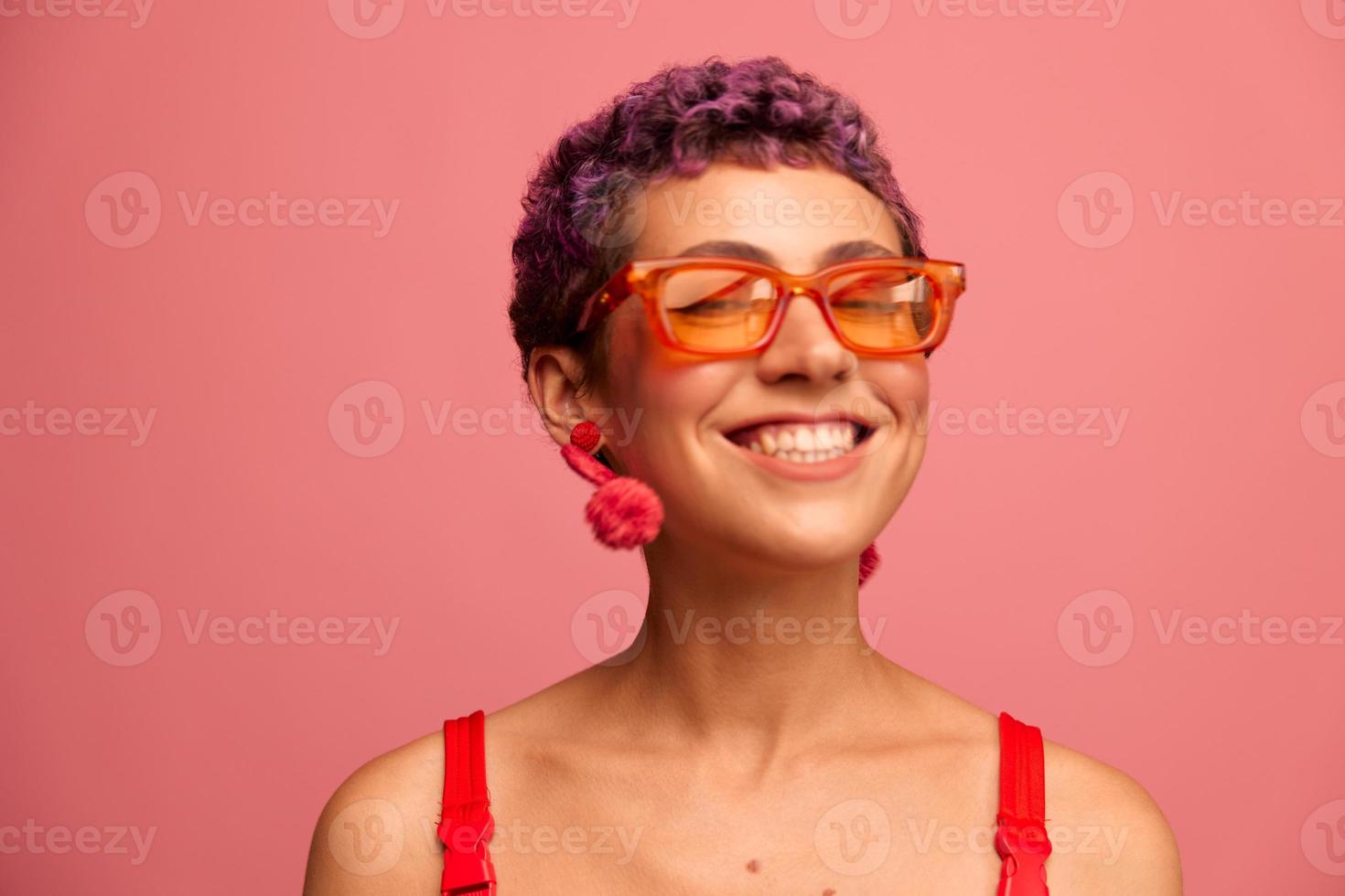moda retrato do uma mulher com uma curto corte de cabelo dentro colori oculos de sol com incomum acessórios com brincos sorridente em uma Rosa brilhante fundo foto