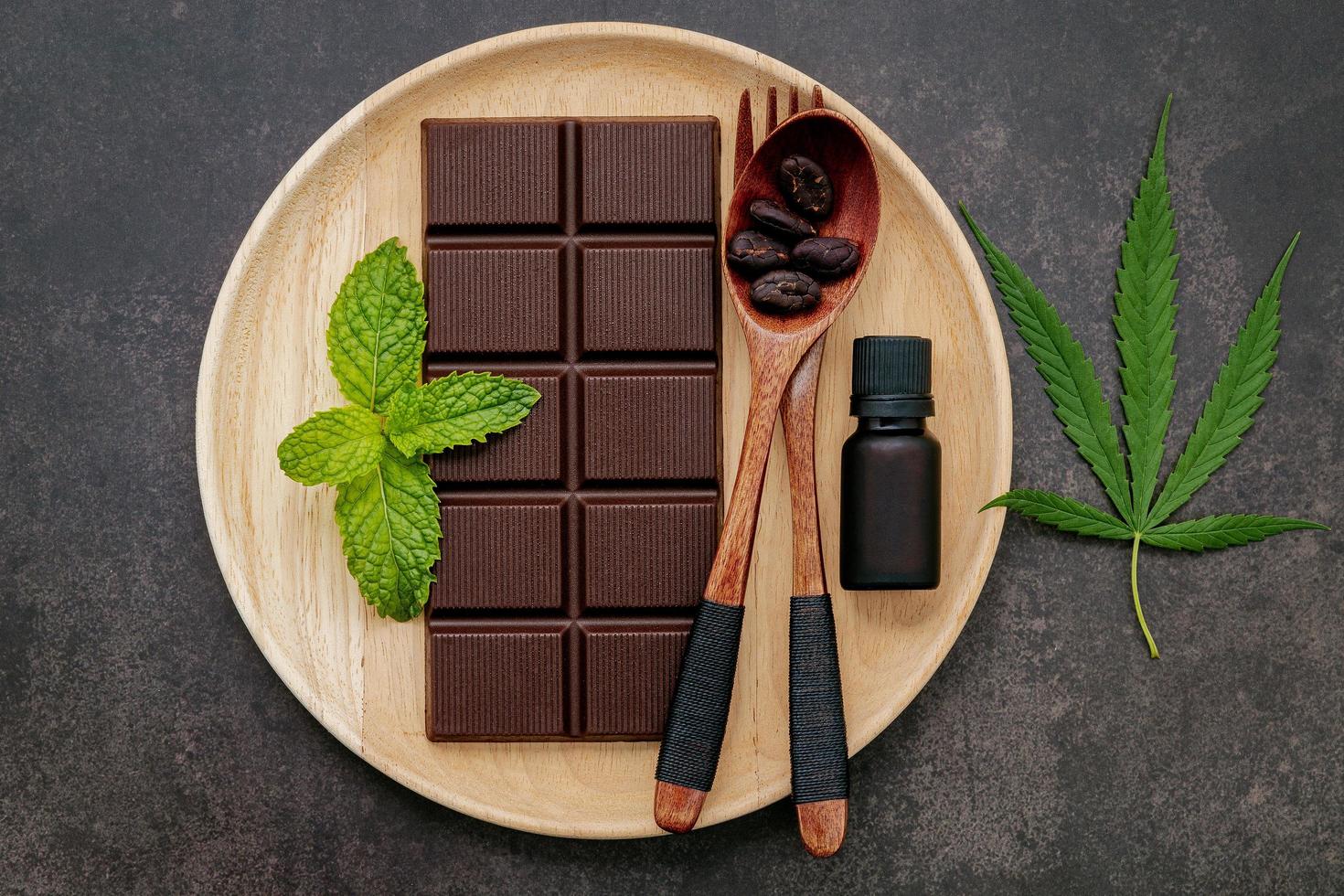 folha de cannabis com chocolate amargo, folhas de plantas e utensílios de madeira em um fundo escuro de concreto foto