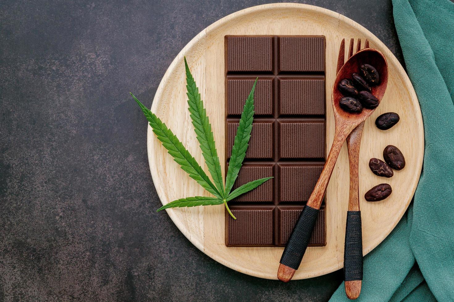 folha de cannabis com chocolate amargo, folhas de plantas e utensílios de madeira em um fundo escuro de concreto foto