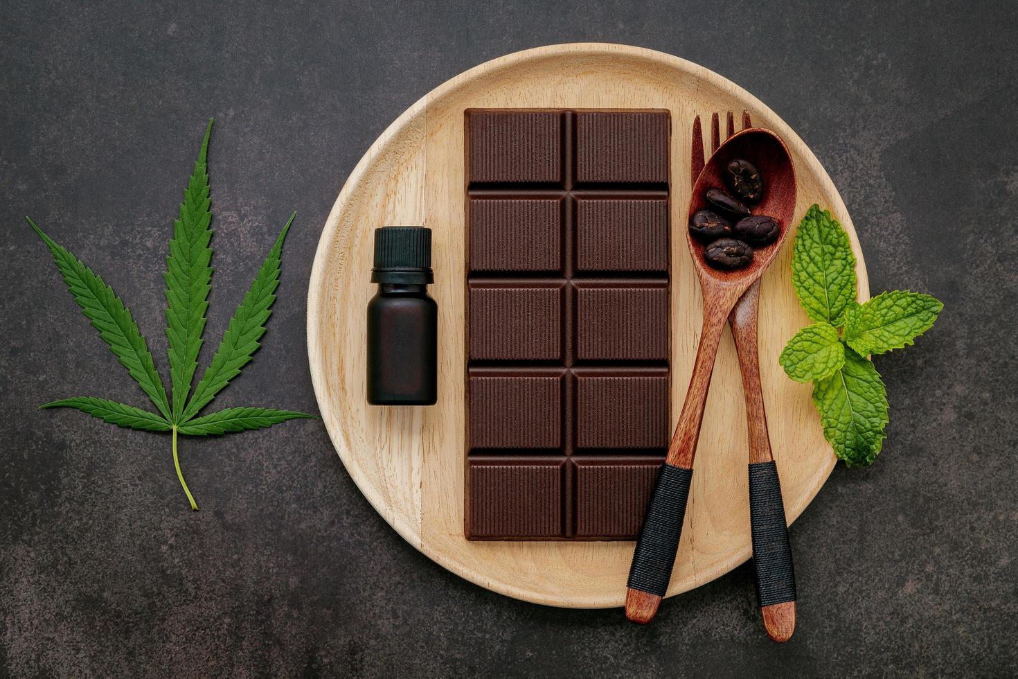 folha de cannabis com chocolate amargo, folhas de plantas e utensílios de madeira em um fundo escuro de concreto foto