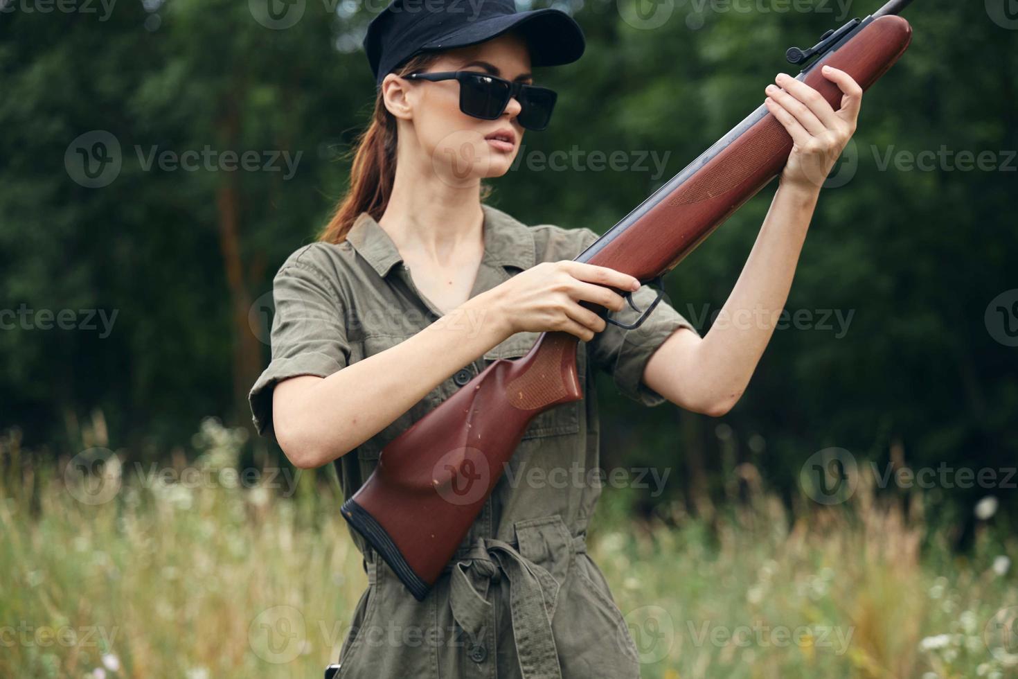 mulher em natureza detém uma arma dentro dele mãos, oculos de sol, verde macacão Preto boné foto