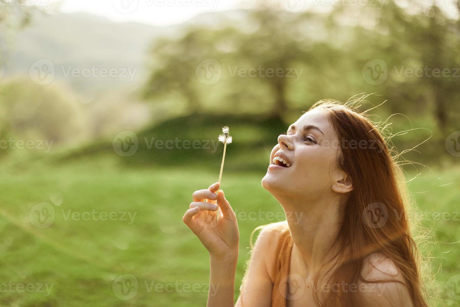 a feliz mulher sorrisos e golpes a dente de leão dentro a vento. verão verde panorama e brilho do sol dentro a fundo foto