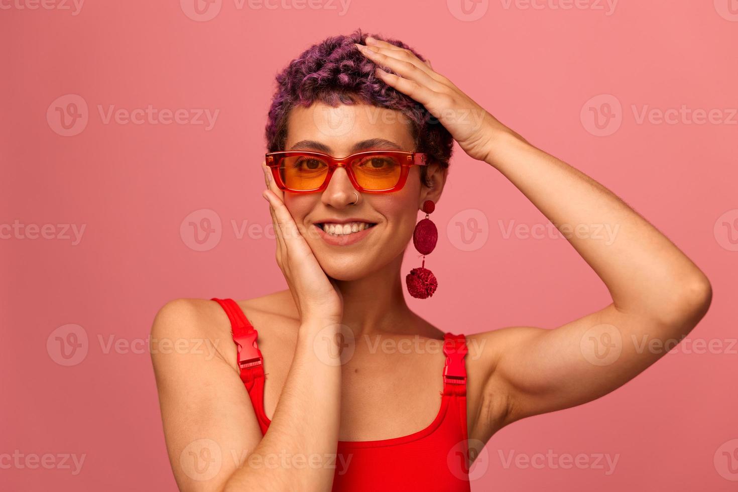 moda retrato do uma mulher com uma curto corte de cabelo dentro colori oculos de sol com incomum acessórios com brincos sorridente em uma Rosa brilhante fundo foto