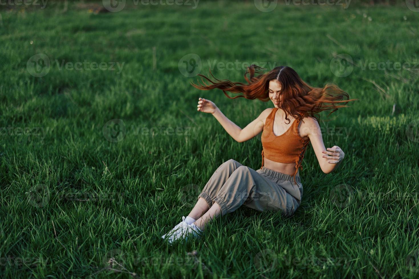 uma ruivo mulher com longo, varrido pelo vento cabelo senta ao ar livre em a Relva dentro a parque e sorrisos, a pôr do sol luz iluminador dela face. a conceito do harmonia com natureza ao ar livre foto