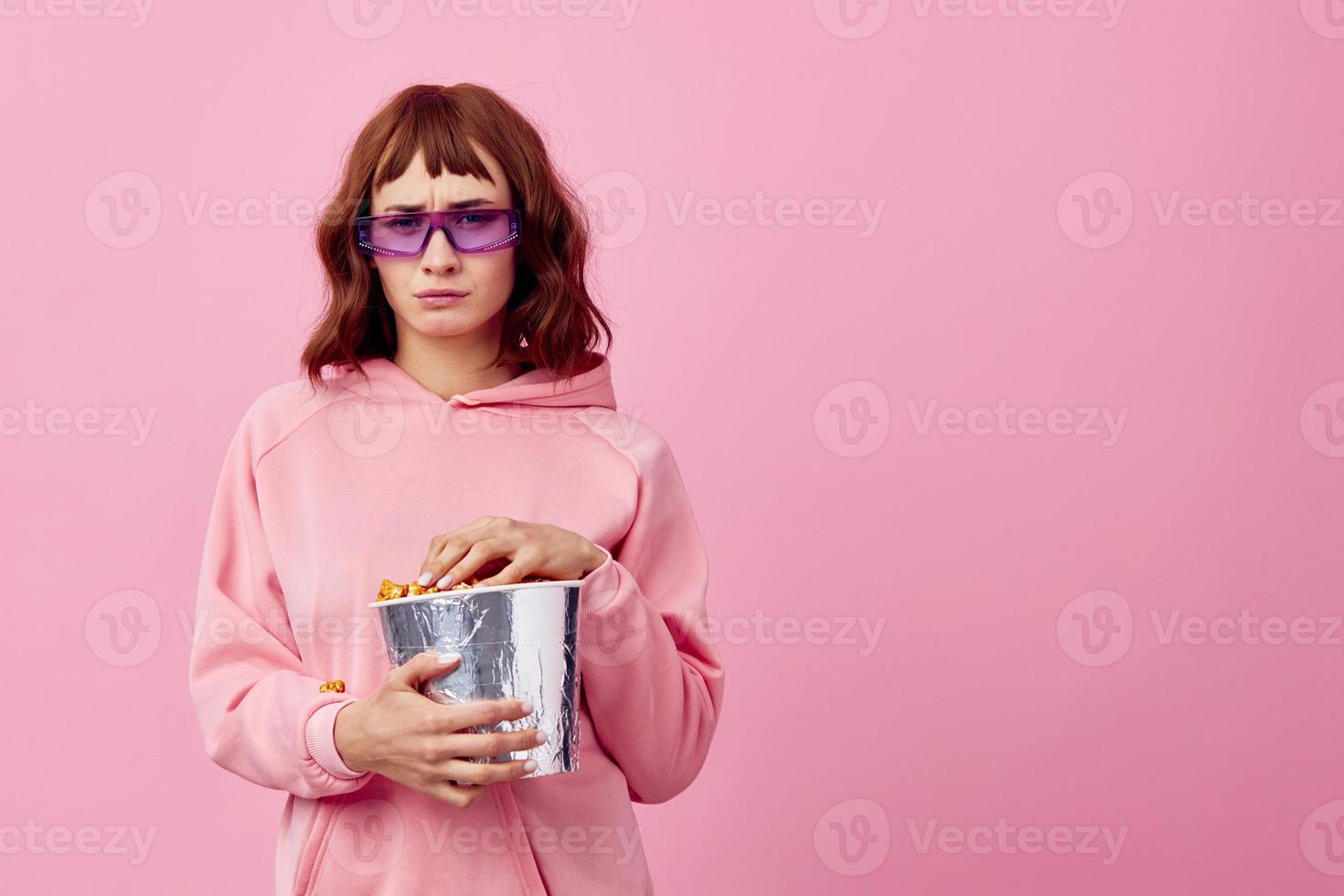 legal filme. confuso fofa ruiva senhora dentro Rosa moletom com capuz oculos de sol com Pipoca posando isolado em sobre Rosa estúdio fundo mostrar cópia de espaço bandeira. moda cinema conceito. entretenimento oferta foto