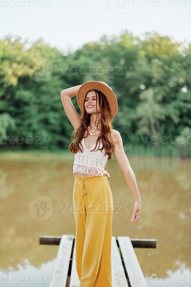 uma jovem mulher dentro uma hippie Veja e vestido ecológico viaja dentro natureza de a lago vestindo uma chapéu e amarelo calça dentro a outono pôr do sol foto