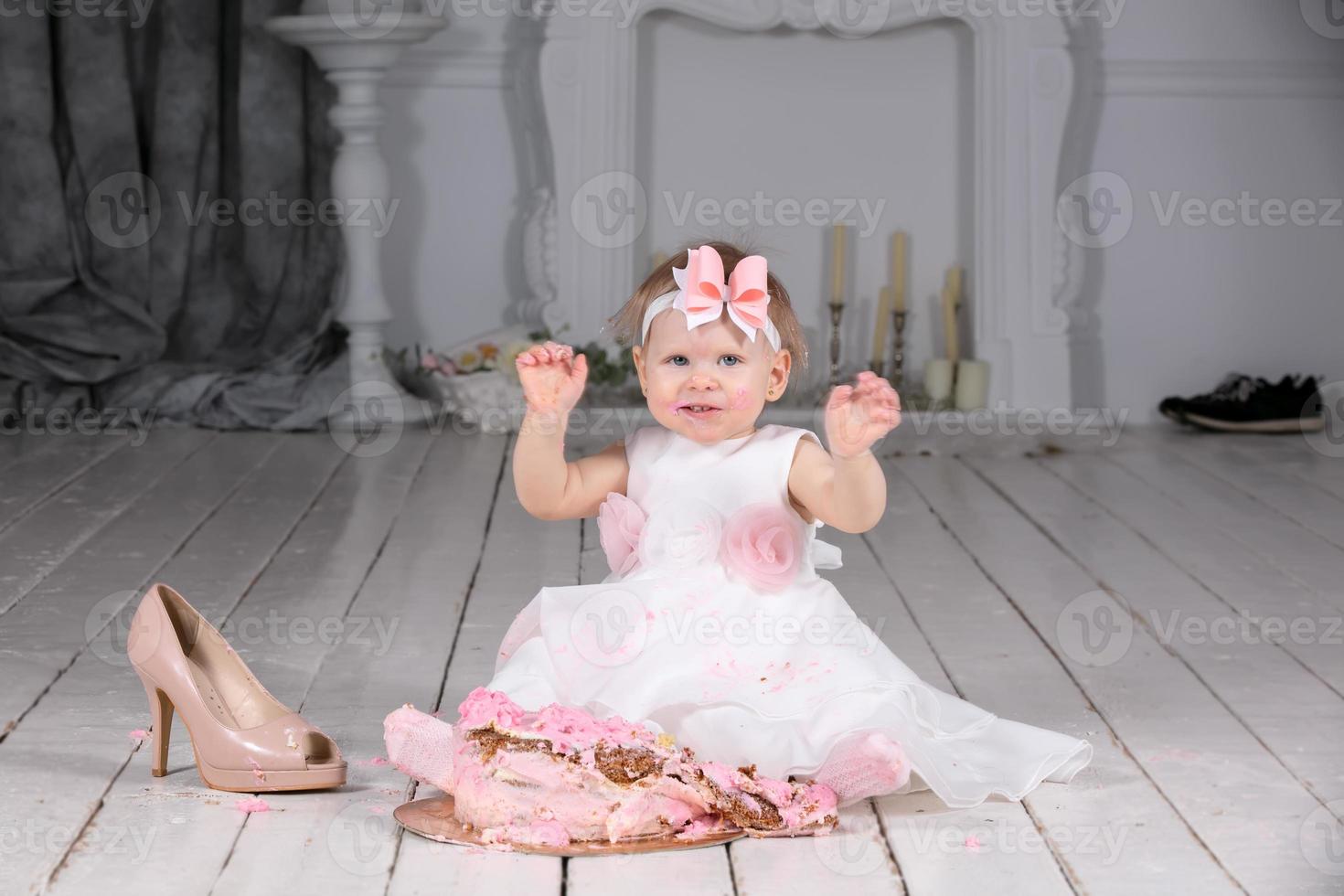 pequeno engraçado anos menina com uma festivo bolo, manchado com creme, é sentado em a chão. foto