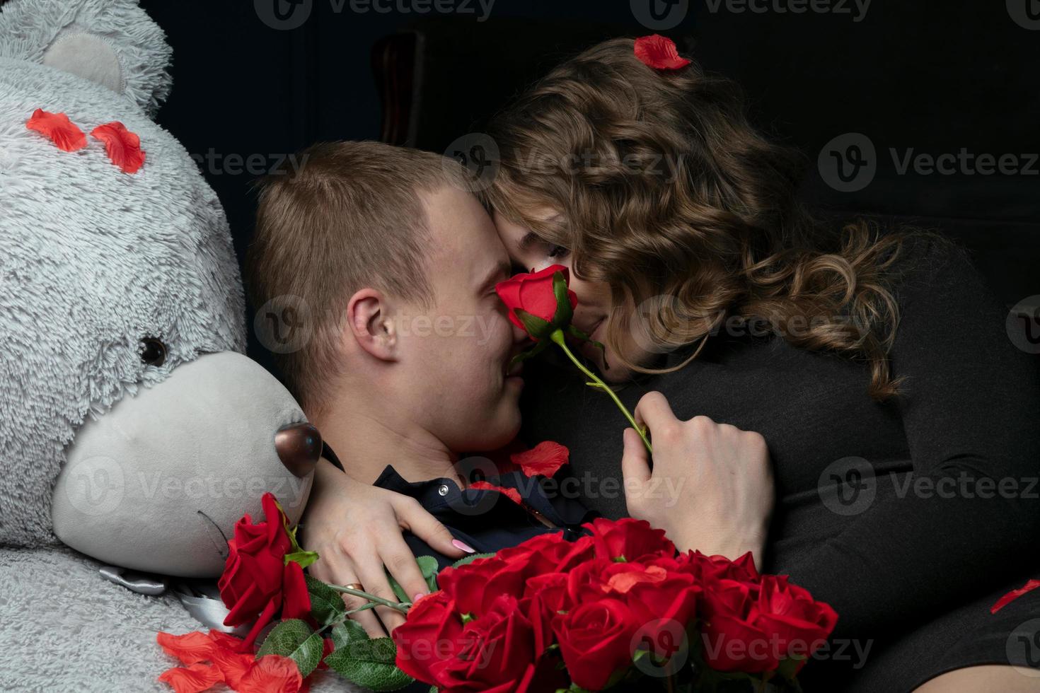 lindo jovem casal às lar. abraços, Beijos e goza gastos Tempo junto, a comemorar dia dos namorados dia com vermelho rosas em uma Urso de pelúcia urso. foto