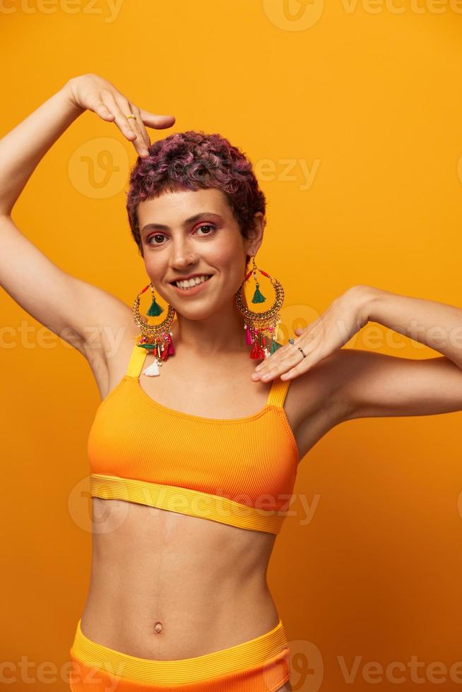 retrato do uma jovem mulher com uma curto corte de cabelo e colori cabelo sorridente dentro amarelo e mostrando dela língua às a Câmera em a laranja fundo dançando com brincos acessórios dentro a estúdio foto