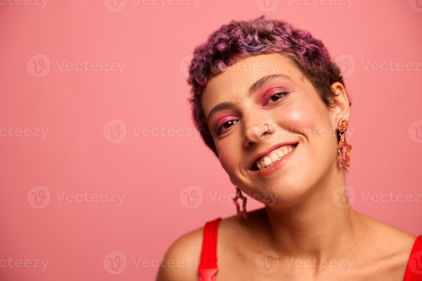 moda retrato do uma mulher com uma curto corte de cabelo do roxa cor e uma sorrir com dentes dentro uma vermelho topo em uma Rosa fundo dançando alegremente foto