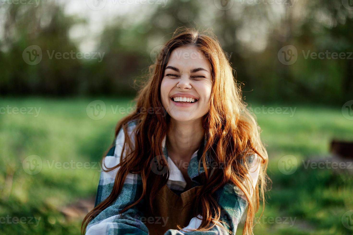 engraçado mulher retrato fechar-se rindo e balançando para a Câmera dentro frente do uma verde parque dentro verão, a pôr do sol brilhando em dela vermelho cabelo foto