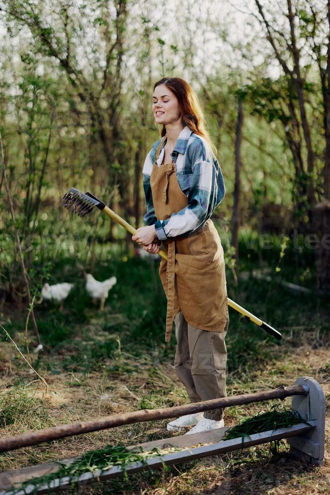 uma mulher sorridente belas e olhando às a Câmera, uma agricultor dentro trabalhos roupas e a avental trabalhando ao ar livre dentro natureza e segurando uma ancinho para reunir Relva e forragem para a animais dentro a jardim foto