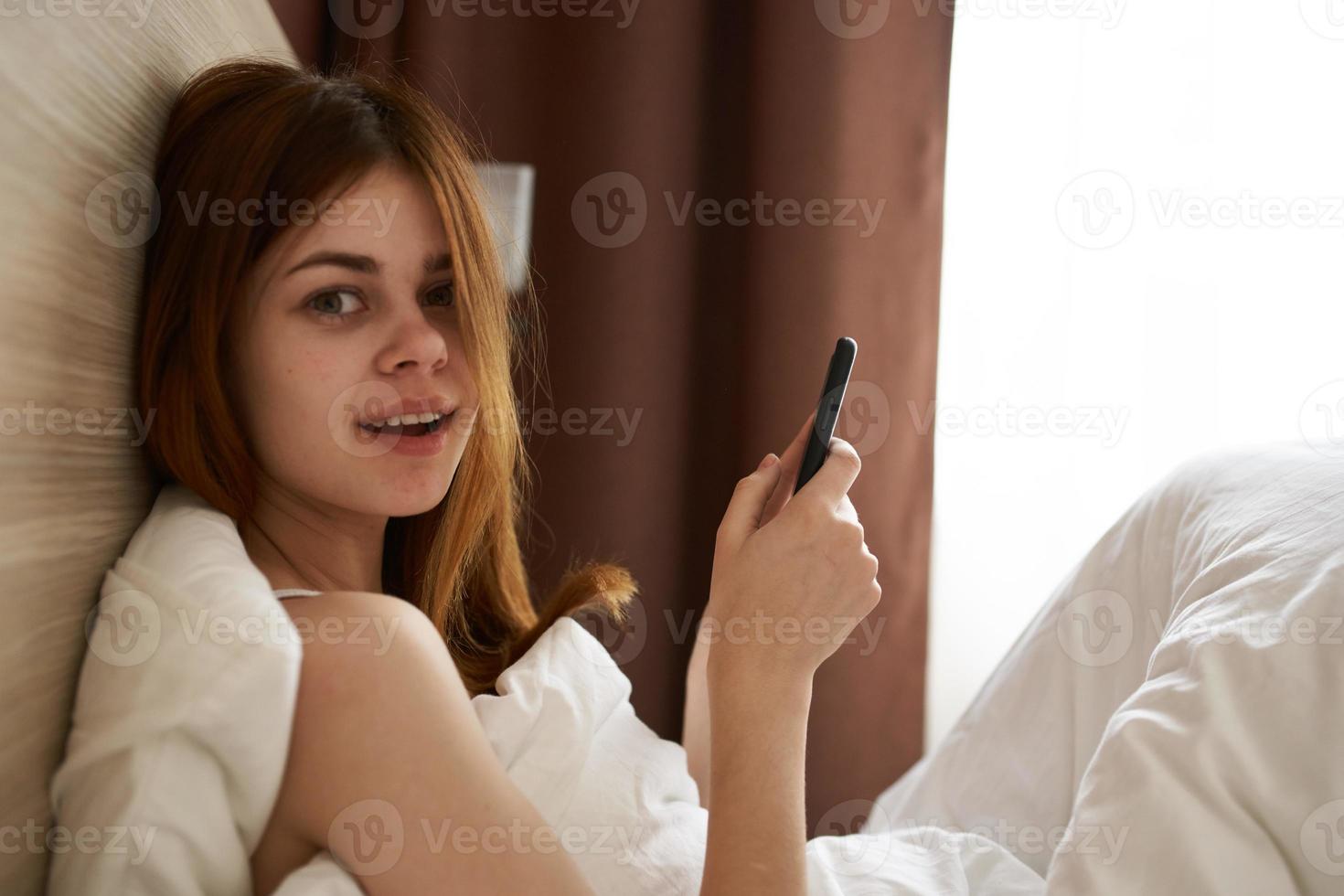 mulher com Móvel telefone mentiras dentro cama e janela cobertor cortinas foto