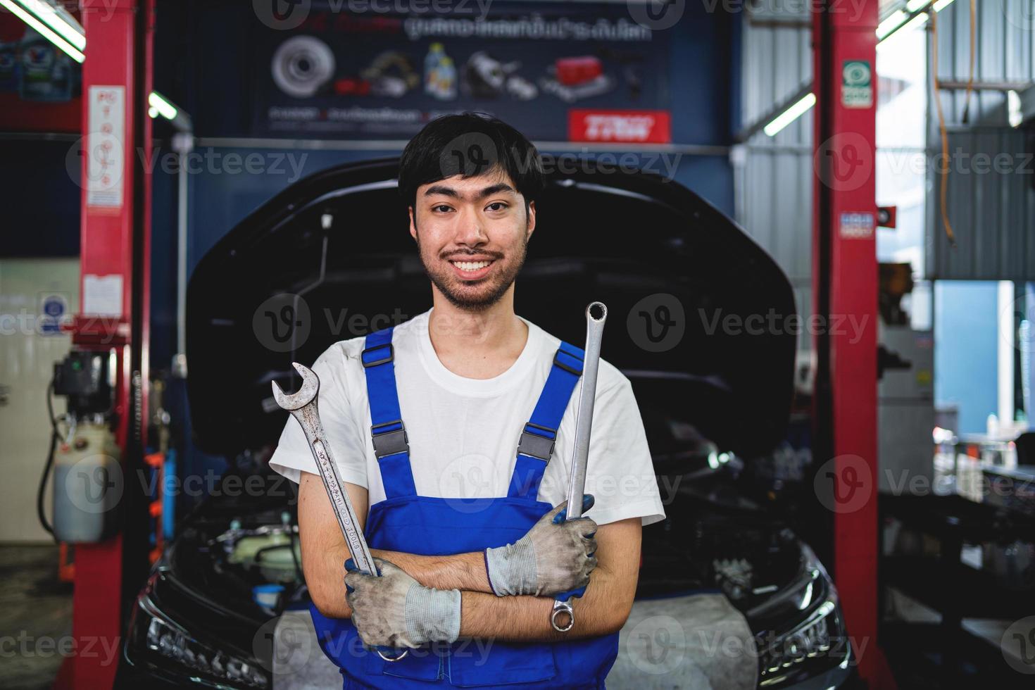 retrato do sorridente ásia masculino reparador segurando de pernas cruzadas chave inglesa às garagem profissional reparar e manutenção. carro inspeção serviço foto