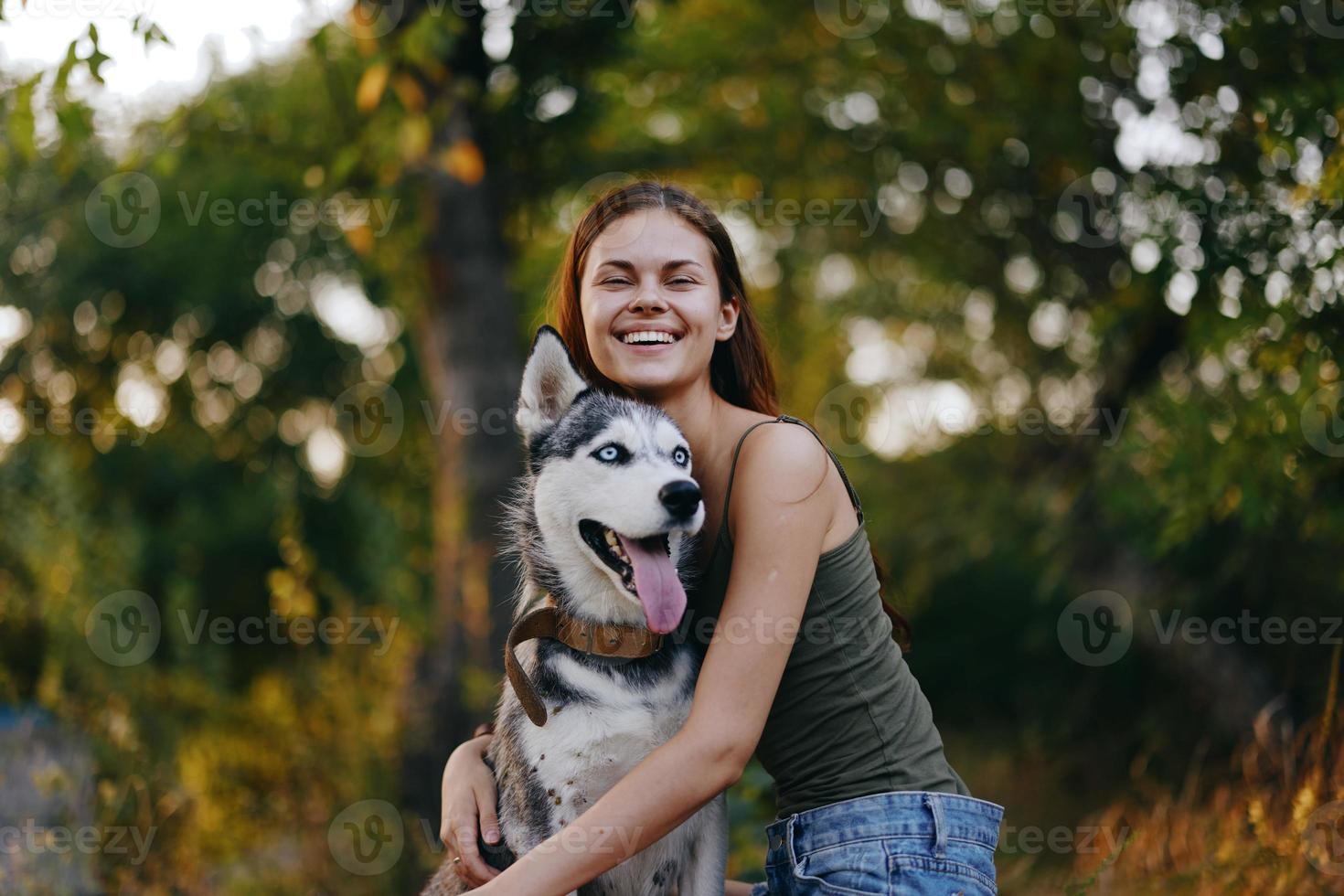 alegre mulher com uma rouco procriar cachorro sorridente enquanto sentado dentro natureza em uma andar com uma cachorro em uma trela outono panorama em a fundo foto