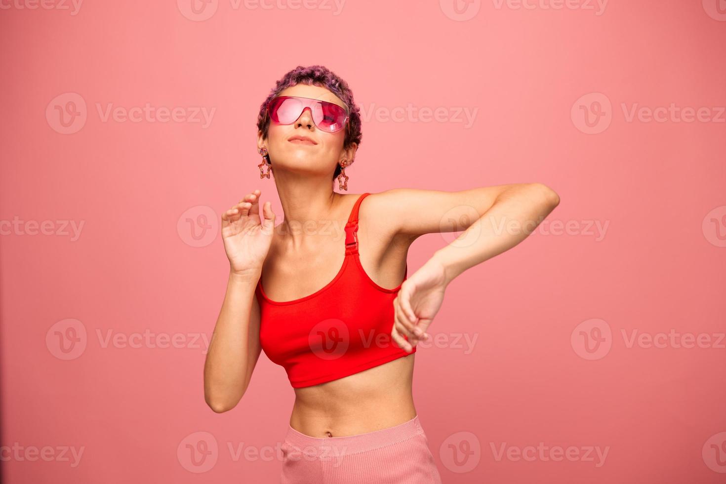 moda retrato do uma mulher com uma curto corte de cabelo dentro colori oculos de sol com incomum acessórios com brincos sorridente em uma Rosa brilhante fundo com uma ginástica corpo dançando foto