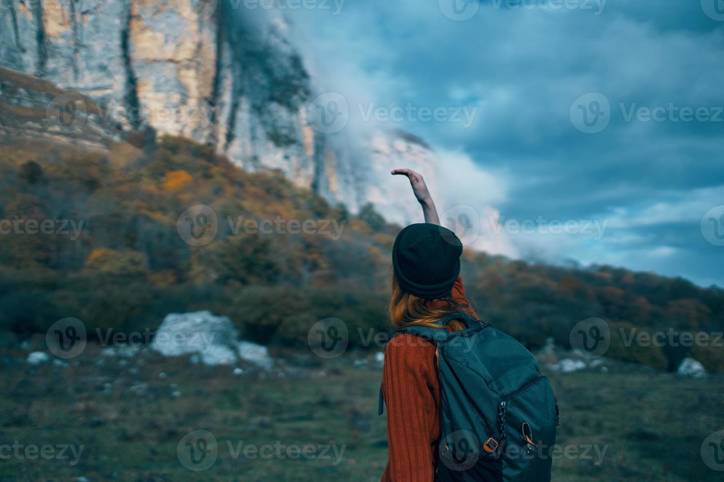 uma viajante com uma mochila e dentro uma vermelho suéter mostra a montanhas com dela mão e a azul céu nuvens foto