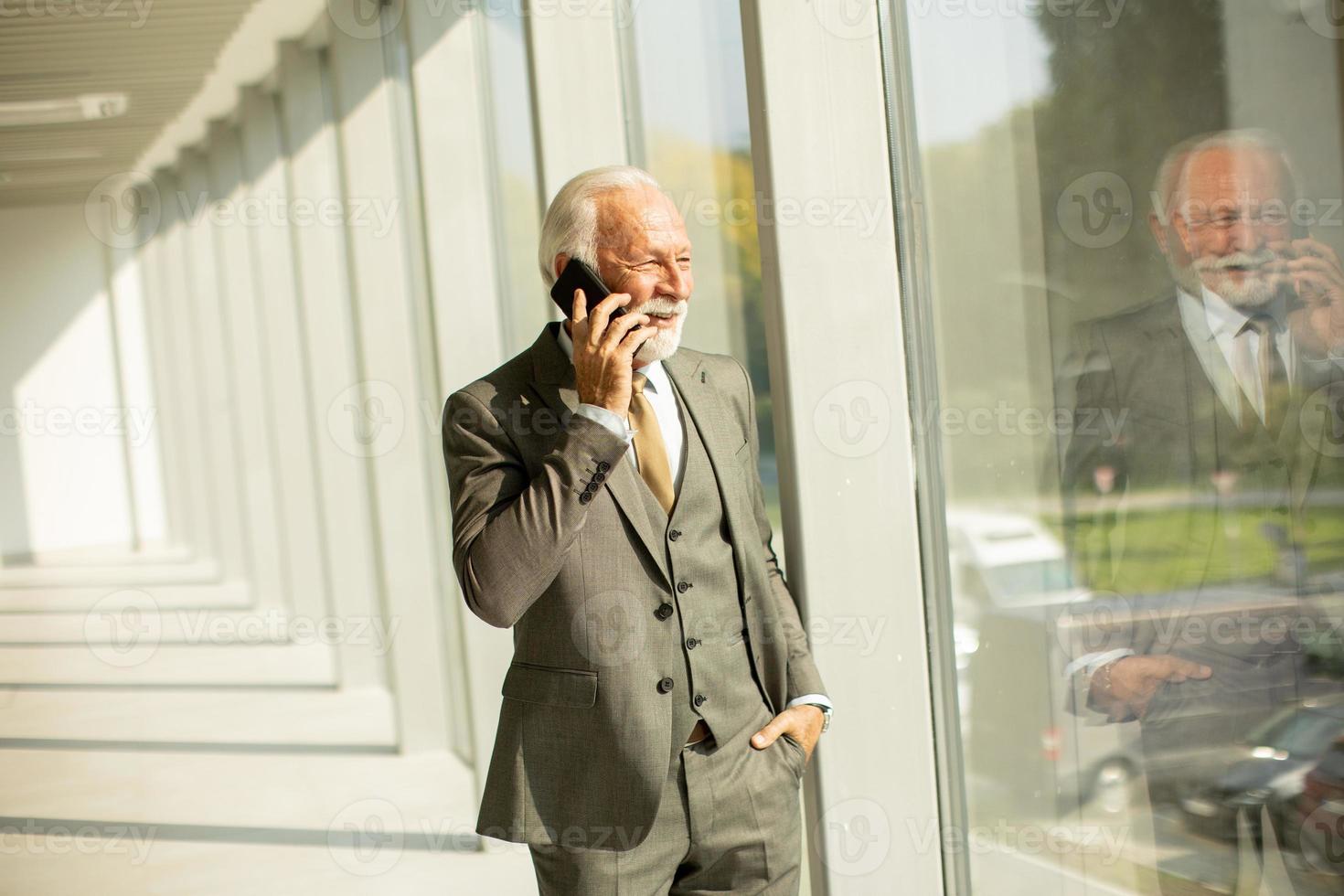 uma Senior o negócio homem carrinhos dentro a escritório corredor, focado em dele Móvel telefone foto