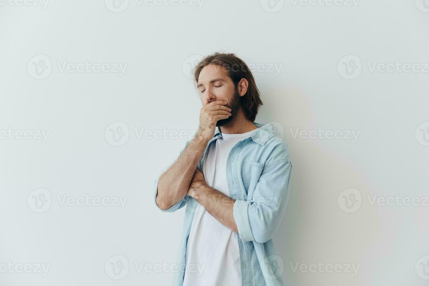 milenar homem com uma barba carrinhos contra uma branco parede e cobre dele face com dele mão a partir de tristeza, estresse foto