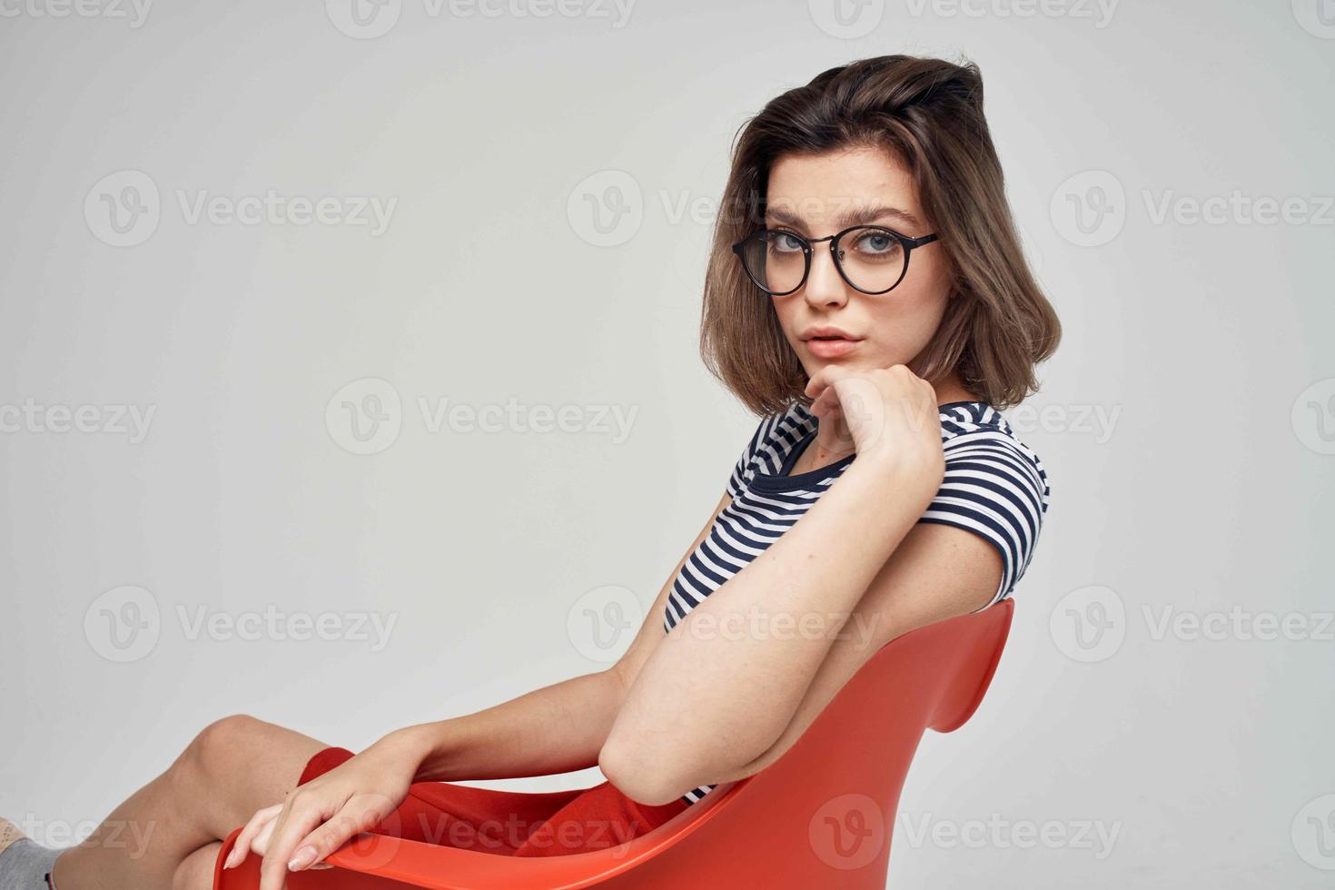 lindo mulher dentro uma listrado camiseta sentado em a vermelho cadeira moderno estilo Penteado foto