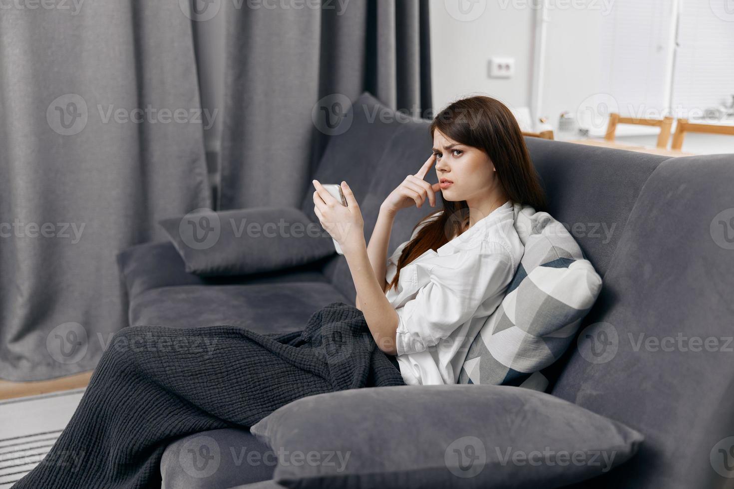 uma mulher dentro uma confortável apartamento com uma Móvel telefone dentro dela mão senta em a sofá foto
