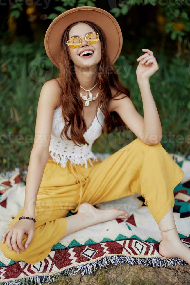 hippie mulher sorridente dentro eco roupas amarelo calça, branco tricotar principal, chapéu e amarelo óculos sentado em xadrez dentro parque assistindo pôr do sol, estilo de vida acampamento viagem foto