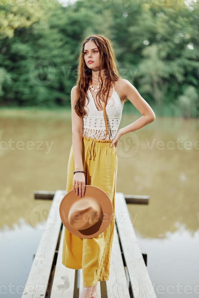 uma jovem mulher dentro uma hippie Veja e vestido ecológico dançando ao ar livre de a lago vestindo uma chapéu e amarelo calça dentro a verão pôr do sol foto
