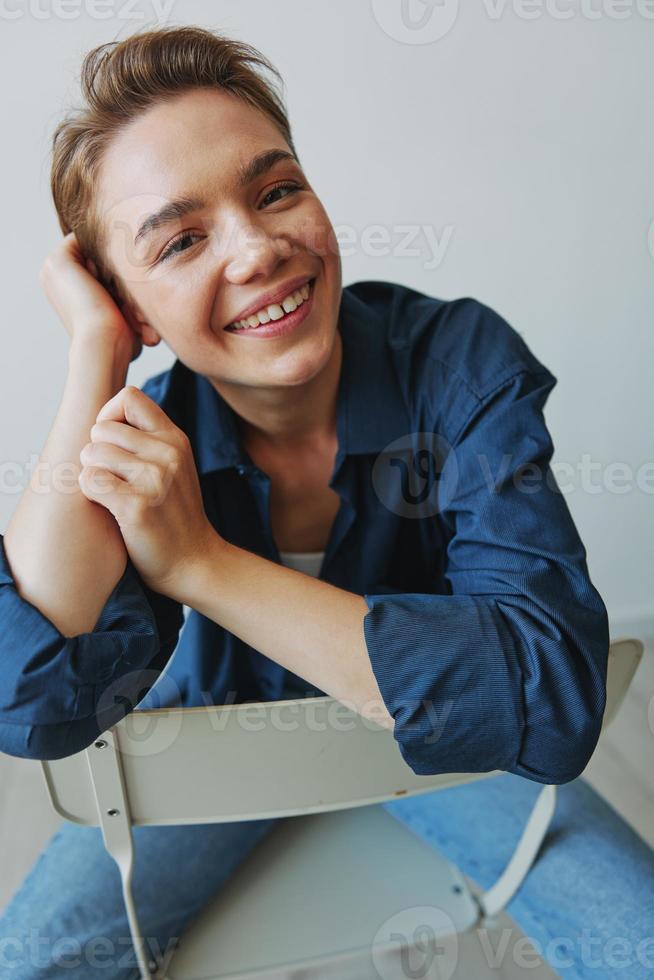 uma jovem mulher sentado dentro uma cadeira às casa sorridente com dentes com uma curto corte de cabelo dentro jeans e uma jeans camisa em uma branco fundo. menina natural poses com não filtros foto