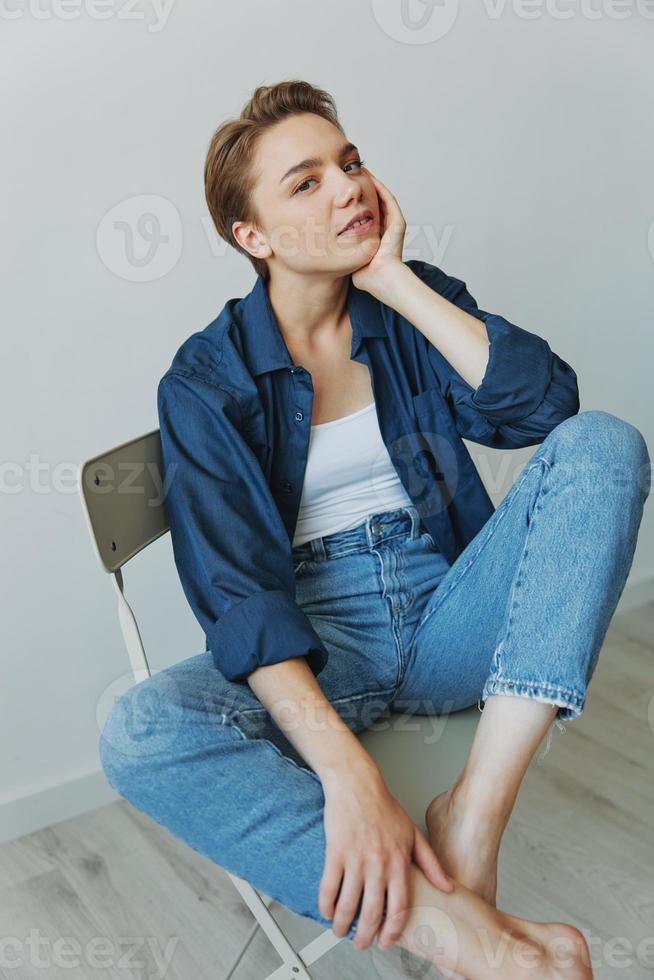 uma jovem mulher sentado dentro uma cadeira às casa sorridente com dentes com uma curto corte de cabelo dentro jeans e uma jeans camisa em uma branco fundo. menina natural poses com não filtros foto