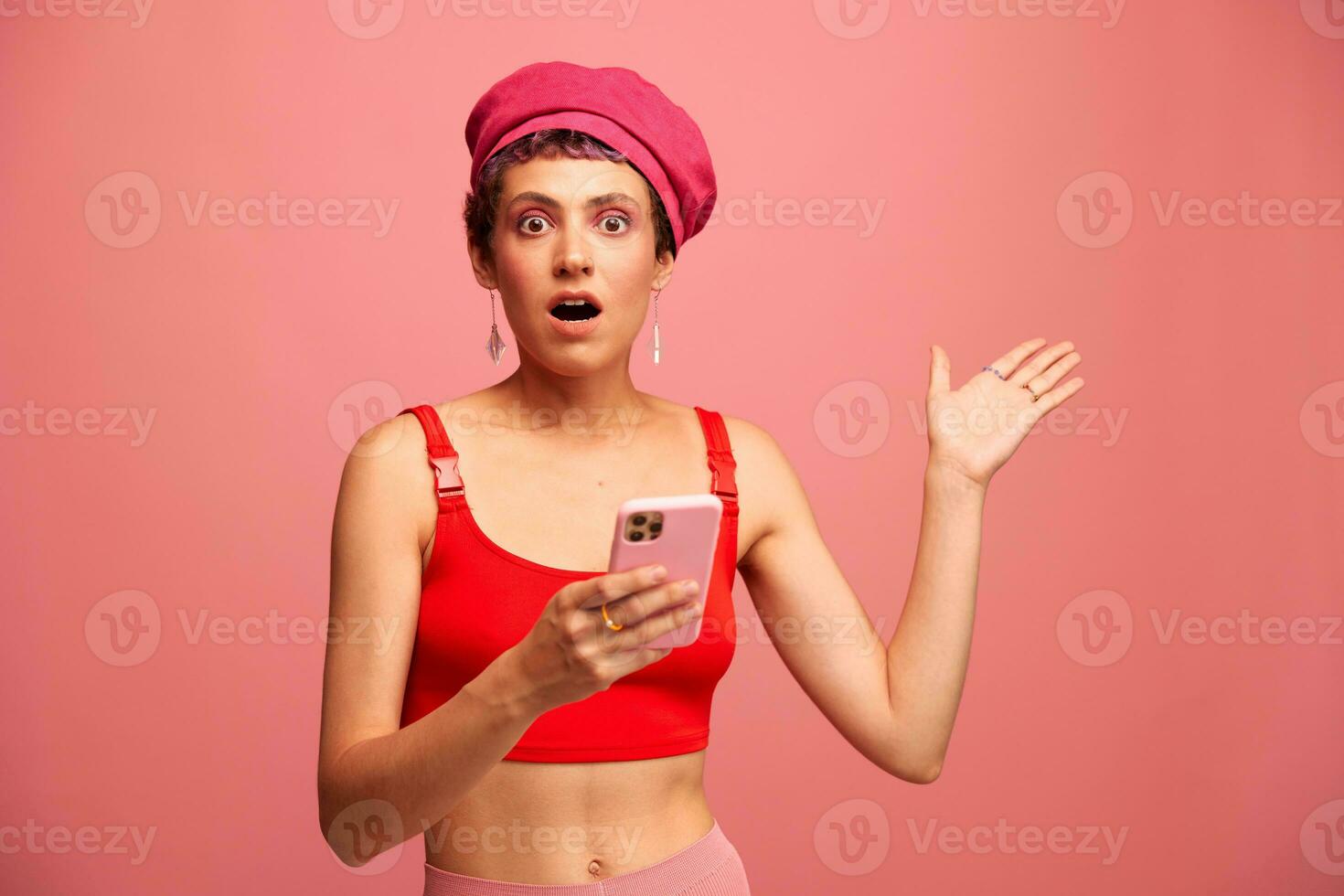 jovem mulher blogueiro com colori Rosa cabelo e curto corte de cabelo lançando através a telefone tela e digitando uma mensagem com uma sorrir e surpresa dentro à moda roupas em uma Rosa fundo foto
