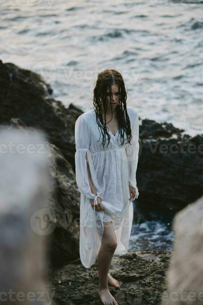 bonita mulher dentro branco vestir carrinhos em a pedras oceano natureza liberdade foto