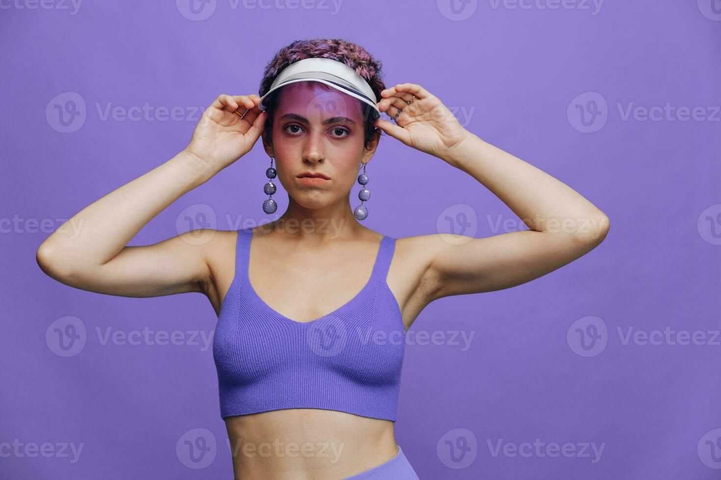 retrato do uma desportivo moda mulher posando dentro uma roxa Esportes terno para ioga e uma transparente boné em uma roxa fundo monocromático foto