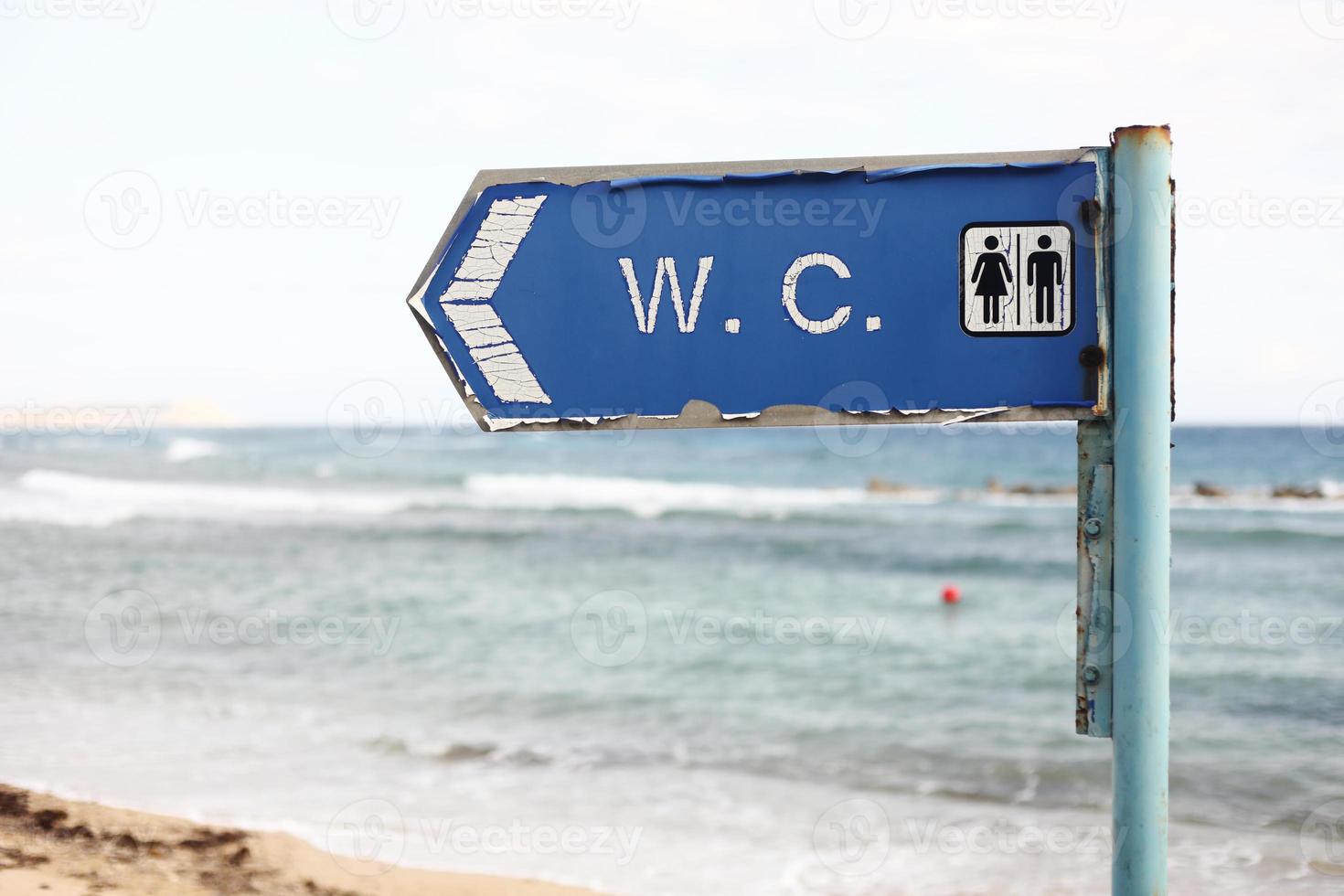 direção ao banheiro em uma praia. inscrição wc na placa de banheiro pendurada em um poste com um fundo do mar e do céu foto