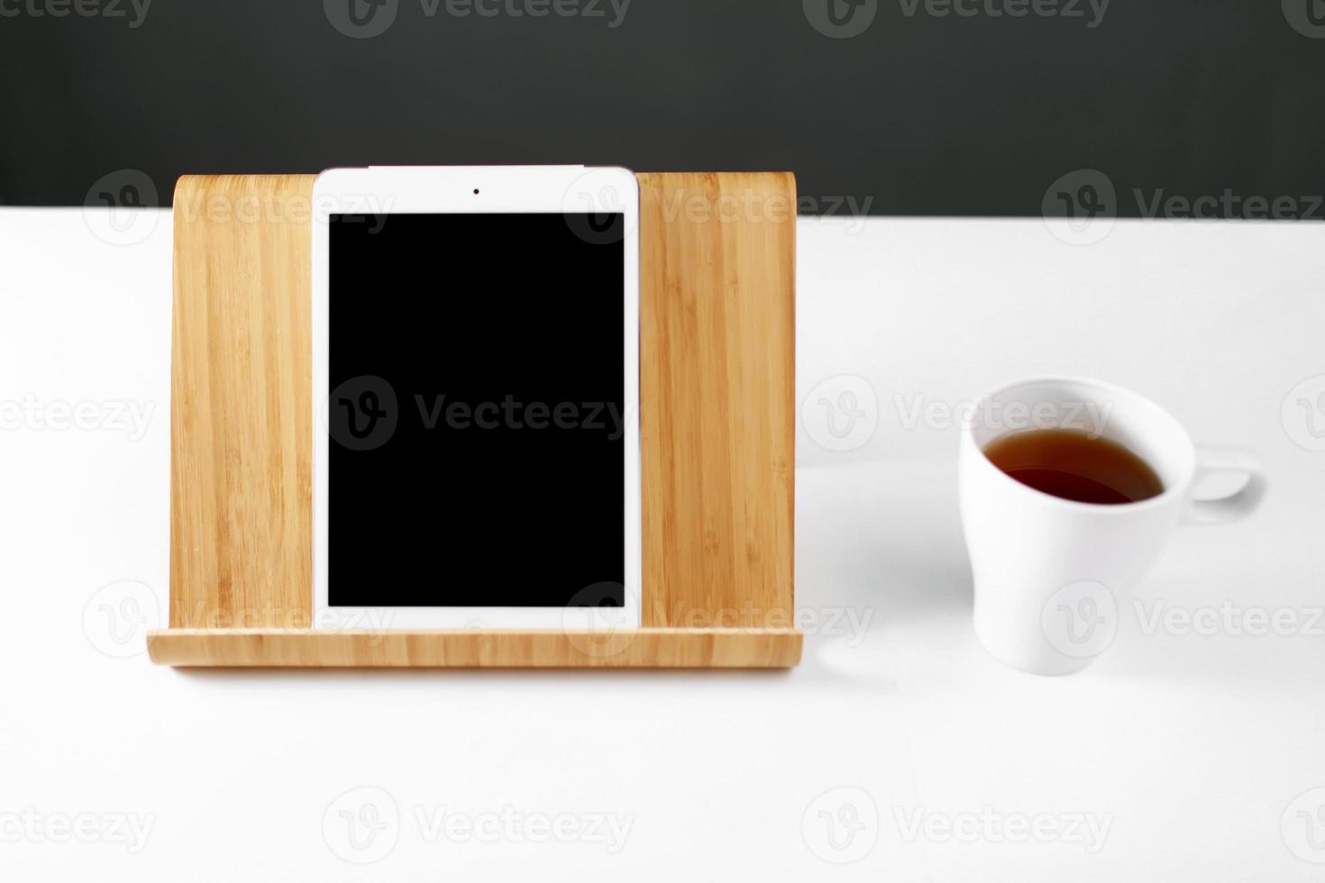 tablet no suporte. caneca branca com chá. tablet digital de maquete em um suporte de madeira. local de trabalho de escritório foto