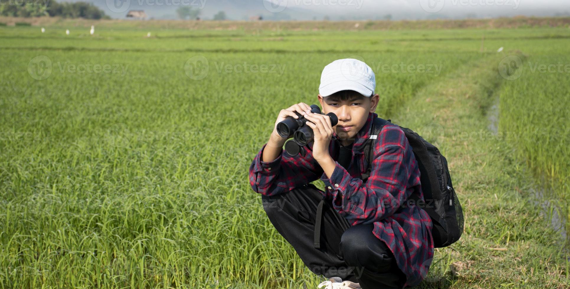 ásia Garoto vestindo xadrez camisa e uma boné segurando uma binóculos e olhando às a rebanho do pássaros alimentando em a arroz campo e vôo dentro a céu dentro a arroz campo. foto
