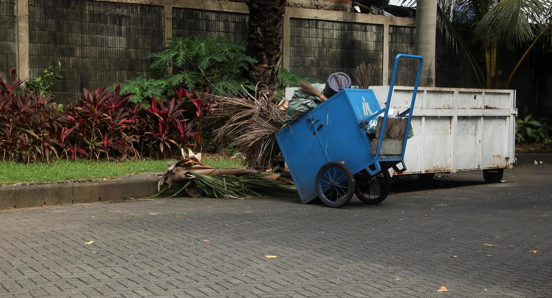 1 azul puxar Lixo carrinho com rodas. Gerobak sampah biru fotografia objeto ao ar livre isolado em minúsculo jardim e tijolo terra pavimentos fundo. foto