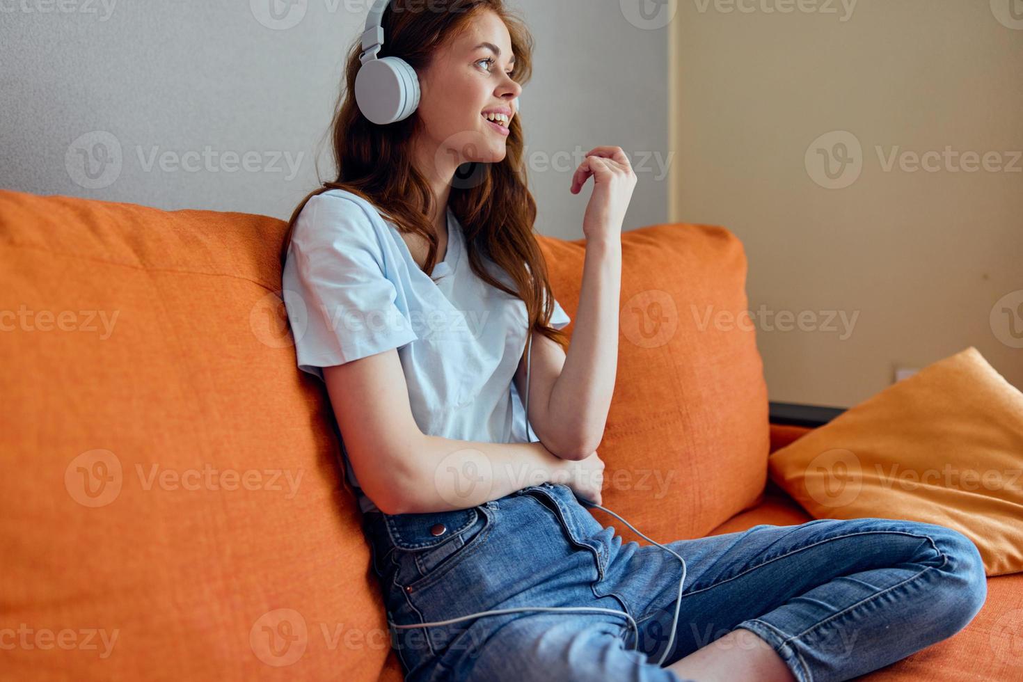 sorridente mulher ouvindo para música com fones de ouvido em a laranja sofá tecnologias foto