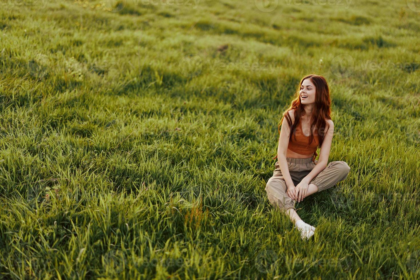 uma ruivo mulher senta dentro natureza em a Relva com dela mãos atrás dela cabeça e sorridente dentro a pôr do sol claro. a conceito do saudável descansar e meditação para saúde foto