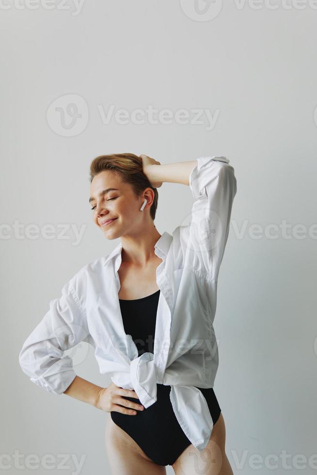 jovem mulher adolescente ouvindo para música com infértil fones de ouvido e dançando lar, sorrindo com dentes com uma curto corte de cabelo dentro uma branco camisa em uma branco fundo. menina natural poses com não filtros foto