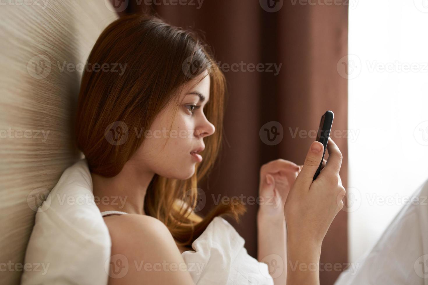 mulher com Móvel telefone mentiras dentro cama e janela cobertor cortinas foto