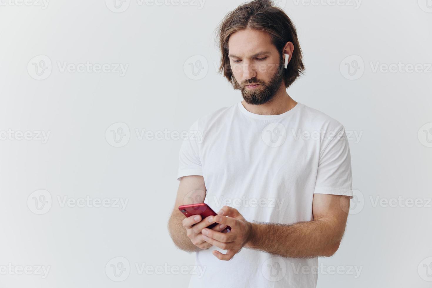 uma homem com uma barba blogueiro dentro uma branco camiseta com uma telefone e sem fio fones de ouvido parece para dentro a telefone e spreads dele mãos para a lado em uma branco fundo isolado cópia de espaço foto