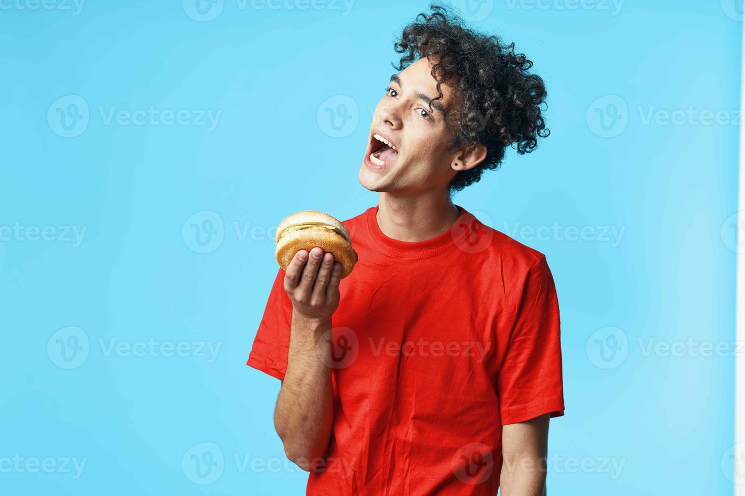 alegre cara com encaracolado cabelo dentro uma vermelho camiseta com uma Hamburger dentro dele mãos velozes Comida dieta foto