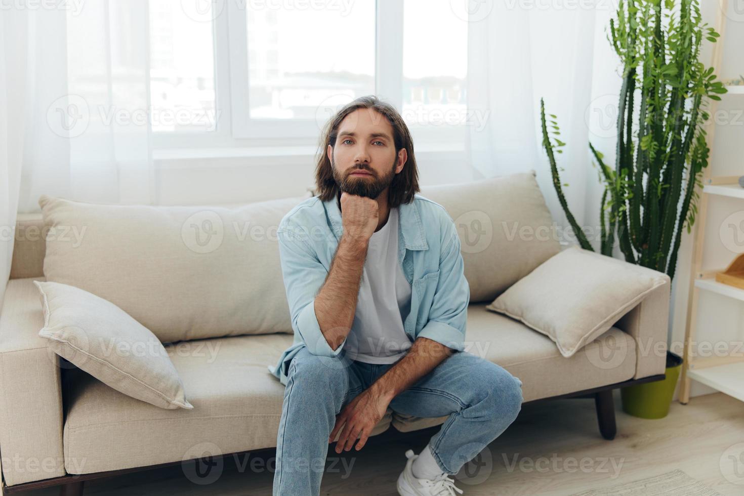 uma freelance homem dentro uma branco camiseta, azul jeans, e camisa senta em a sofá com dele telefone às casa em dele dia fora e tocam jogos foto